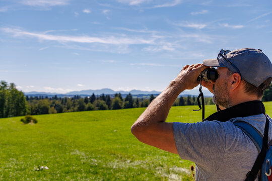 Man with backpack looking through binoculars at alpine mountain panorama across green meadow, nature watching and outdoor exploration concept