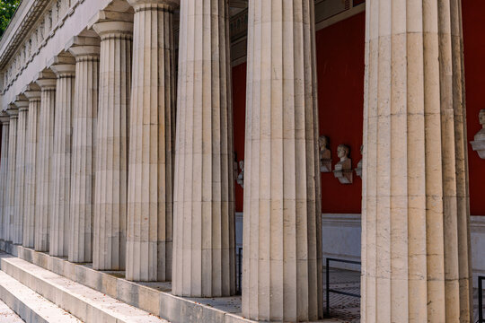 Row of classic neoclassical Doric stone columns and white marble busts along red wall at Ruhmeshalle hall of fame in Munich Bavaria, neoclassical architecture and historic landmark concept