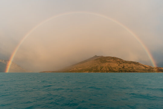 View of a vibrant rainbow arching over the turquoise waters of Lago Argentino with a mountain landscape in the background in El Calafate, Santa Cruz Province, Argentina.