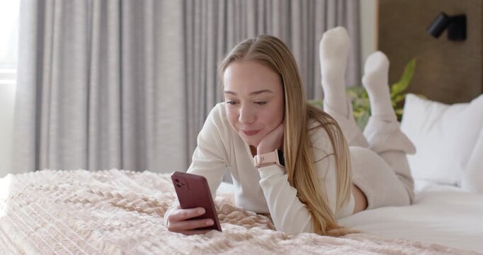 Woman in twenties lying on bed in softly lit bedroom, reading pink phone message, smiling