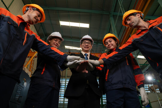 Diverse team of factory workers and manager in hard hats and safety uniforms joining hands together in an industrial manufacturing plant, teamwork, unity and cooperation concept