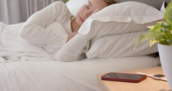 Adult woman opening eyes, reaching and picking up red-cased smartphone on nightstand