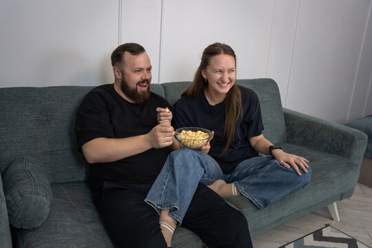 Cheerful couple on a cozy sofa sharing a bowl of popcorn, laughing and relaxing during a casual movie night at home, enjoying a comfortable, intimate evening together