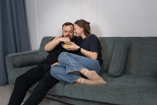 Couple on a sofa in a cozy living room sharing a bowl of popcorn, relaxed and expressive while watching a movie together, enjoying casual home bonding time and comfort
