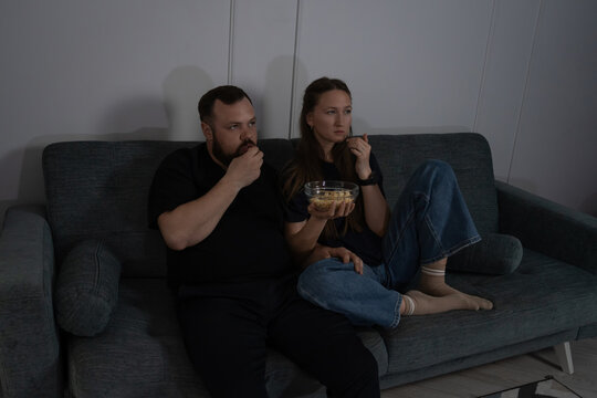 Couple relaxing on a couch in a dark room watching television, enjoying snacks, and spending an evening together in a casual and comfortable home setting