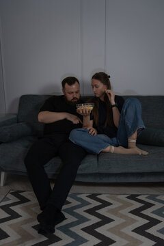 Young couple cuddled on a comfortable sofa in a dim living room, enjoying a cozy movie night with popcorn and snacks, relaxed and spending quality time together indoors. Vertical photo