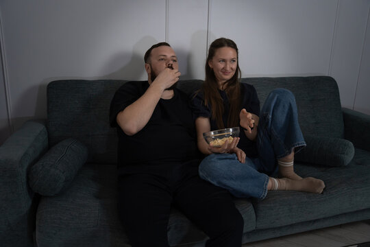 Couple sitting on couch in dimly lit room, sharing snacks and immersed in watching TV, while woman smiling and discussing content with her partner