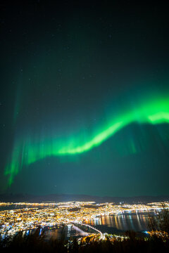 View of the northern lights illuminating the night sky above the city lights and bridge of Tromso, Troms, Norway.