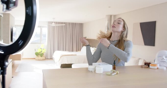 Woman fixing hair, reaching for skincare on table in bedroom, demonstrating gua sha for ring light