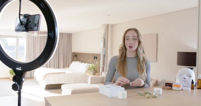 Female clasping hands, speaking to phone near ring light, demoing green facial tool at counter