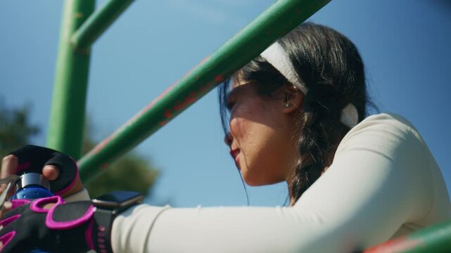 A fit young Asian woman pauses her outdoor exercise routine. She is seen resting on monkey bars and drinking water from a bottle.