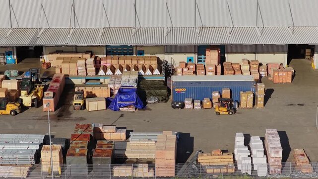 Telephoto aerial view tracking along a sunlit logistics yard in the port of Hamburg, Germany, forklifts among stacks of wooden crates at warehouse loading docks