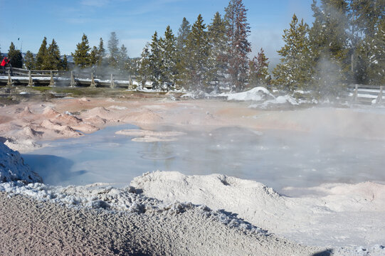 View of a steaming geothermal pool surrounded by snow-covered ground and pine trees with a wooden boardwalk in the background at Yellowstone National Park, United States.