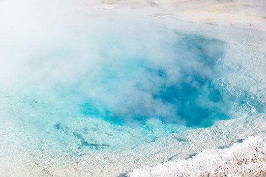 View of a vibrant blue geothermal hot spring with steam rising from the mineral-rich water in Yellowstone National Park, United States.
