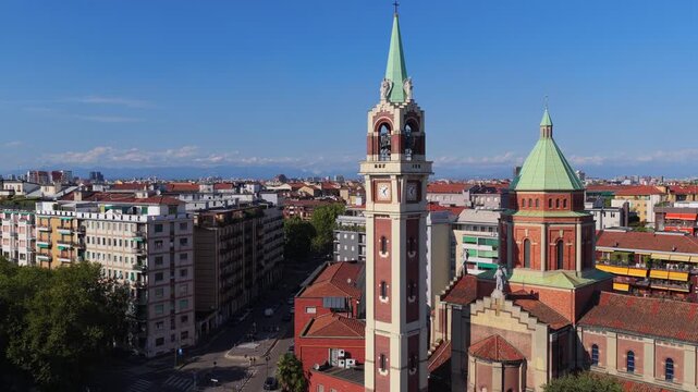 Rising view of brick church with clock bell tower and green copper spires above apartment blocks in Milan, Italy, distant mountains under blue sky