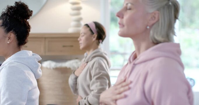 Diverse females following cue, right senior placing hand on heart sensing breath at home by console