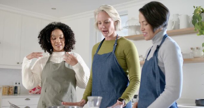 Diverse women fixing aprons after camera pullback, confirming steps, touching mixing bowl at island