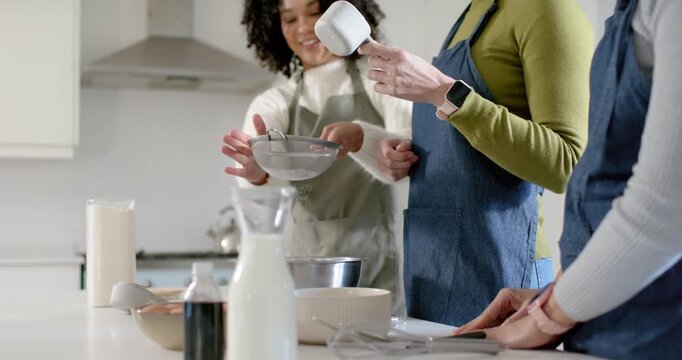 Diverse women in aprons making batter in kitchen, passing measuring cup, sifting sieve