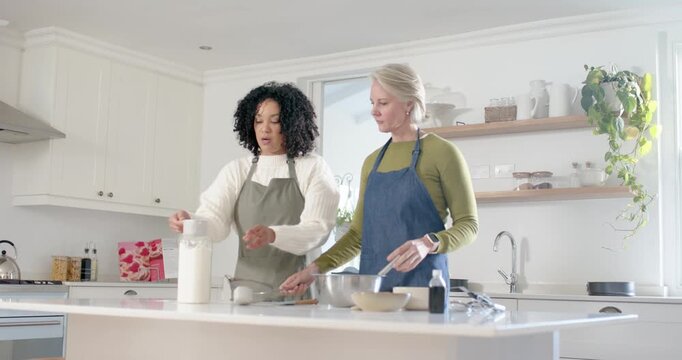 Diverse women reaching for canister measuring and sifting into bowl making batter on kitchen island
