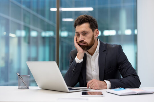 A bored young businessman sits at his desk in the office, looking at his laptop screen
