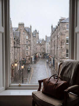 Rainy Edinburgh Street View: A vintage leather travel bag rests on a comfortable armchair by a window, overlooking a cobblestone street in Edinburgh on a rainy day.
