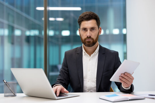 A portrait of a serious young businessman sitting at an office desk, holding a tablet, and looking at the camera