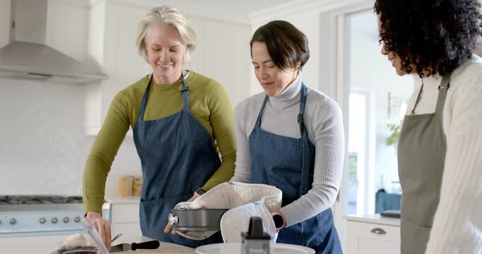 Diverse female friends in kitchen carrying springform pan with oven mitts, inspecting cake doneness