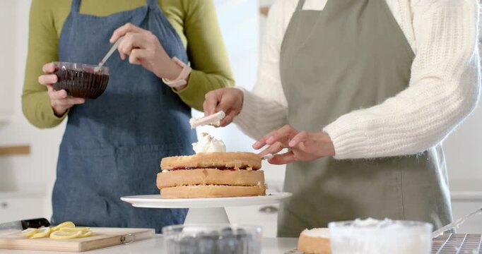 Diverse women reacting to stacked cake, decorating with spatula and stirring jam in kitchen