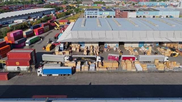 Aerial view flying sideways from colorful container stacks along a busy port warehouse in Hamburg, Germany, trucks and forklifts handling wooden crates, sunny day