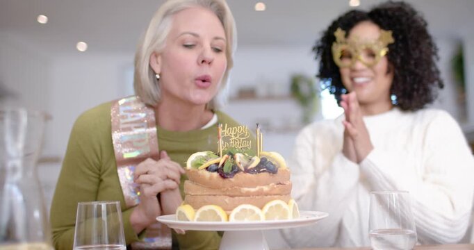 Diverse women clapping, senior blowing lit candles on cake at table with birthday sash, copy space