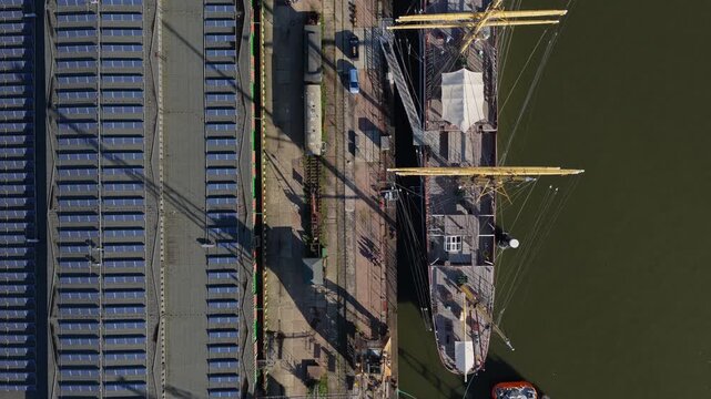 Top down view flying along the historic four-masted barque Peking from stern to bow, yellow masts and yards over dark harbor water beside a quay in Hamburg, Germany