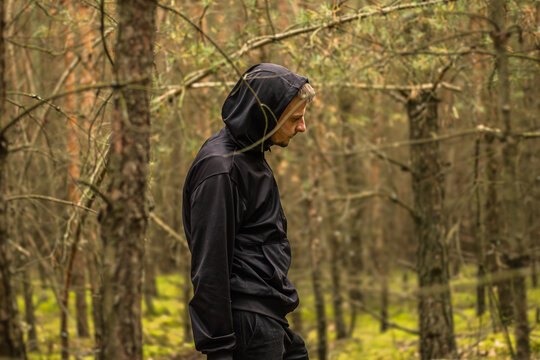 Hooded man in black outdoor clothing standing among slender trees and mossy forest floor, contemplative profile portrait in a woodland setting