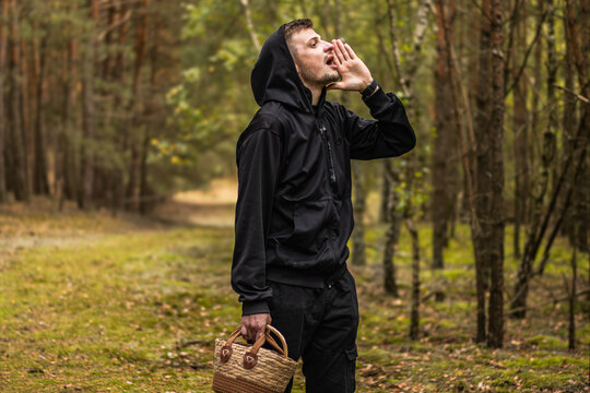 Man in black hooded clothing calling out while carrying a wicker basket along a forest path surrounded by tall trees and mossy ground