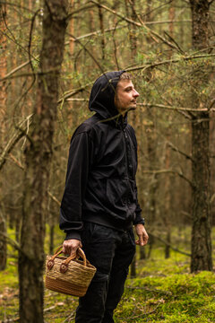 Hooded man carrying a woven basket through a mossy conifer forest with rugged trees and autumn foliage, vertical outdoor portrait in woodland