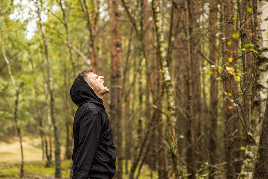 Man in black hooded clothing calling out while carrying a wicker basket along a forest path surrounded by tall trees and mossy ground