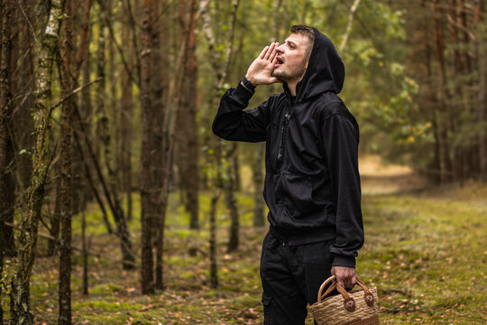 Man in black hooded clothing calling out while carrying a woven basket on a forest trail, surrounded by tall trees and green woodland vegetation