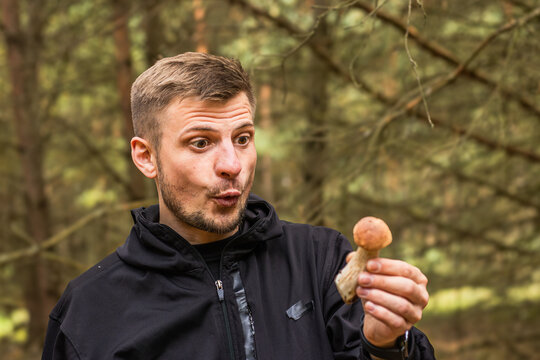 Surprised man holding a small mushroom while porcini examining its unusual shape in a wooded forest setting, outdoor nature portrait with expressive reaction and earthy background