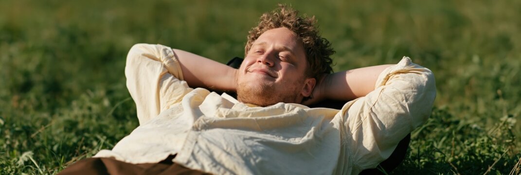 Man lying on grass with eyes closed soaking sunlight for outdoor wellness background and relaxation banner design in natural green meadow