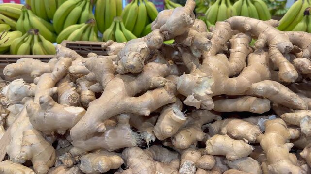 Ginger fills foreground produce stand, piled beneath rows of green bananas. Aromatic root appears fresh and unpeeled, its clustered shapes creating richly textured market display for shoppers.