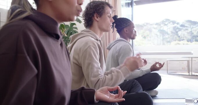 Diverse group starting session, sitting on mats by window in hoodies, shifting hands into mudra