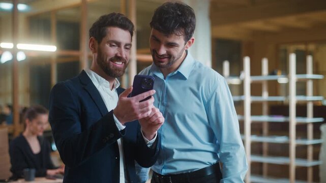 Two happy optimistic Caucasian business men with mobile phone laughing watching humorous video funny concent together in office. Positive businessmen male workers partners laugh look in smartphone