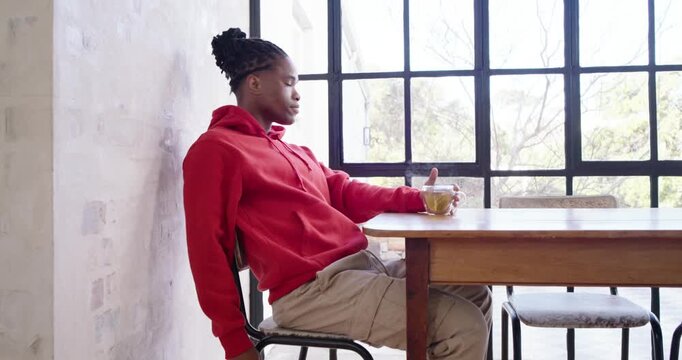 Adult man in red hoodie sitting at dining table with mug, noticing camera, smiling, copy space