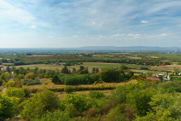 Fototapeta premium Vast panoramic landscape view of rural fields, woodland patches, and distant hills in autumn colors under a bright blue cloudy sky