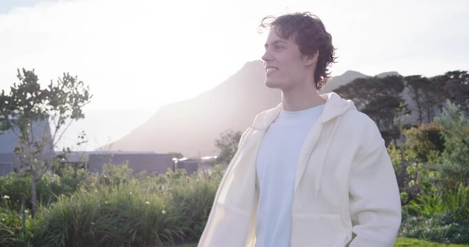 20s man wearing zip hoodie on lawn, wind lifting hair, tucking it, smiling at low sun