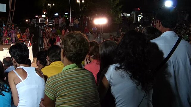 Back view of audience sitting and applauding at an outdoor stage during an evening show