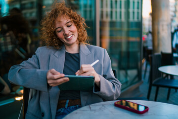 Joyful Caucasian female student operating digital tablet with stylus pen at outdoor cafe, celebrating mobile e-commerce retail store discount codes online shopping sales and offers © BullRun