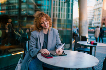Smiling Caucasian female student writing on digital tablet with stylus pen while sitting at outdoor terrace table, checking e-commerce retail app product deals and online discounts © BullRun