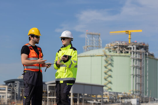 Workers in safety gear check data on tablet near LNG storage facility during day