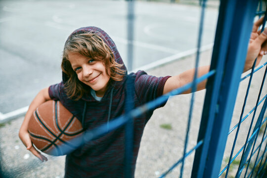 Smiling boy with basketball leaning on outdoor court fence