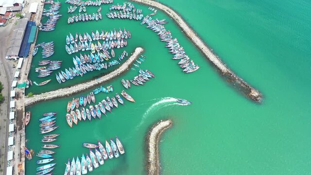 Aerial view of Prigi harbor in Watulimo, featuring numerous colorful fishing boats moored in turquoise waters, protected by stone breakwaters in Watulimo, East Java, Indonesia.